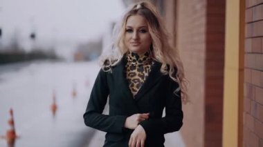 Parlak görünümlü sarışın ceketini düzenler ve ellerini göğsüne geçirir, geniş bir gülümseme sergiler, caddede soyutlanmış, şehir caddesinin bulanık bir köşesinde. Kadın enerjisi. Güzellik
