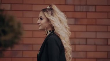 Seksi genç sarışın, profilde, bir tarafa yürür ve kendinden emin bir şekilde bakar, mutlu bir şekilde gülümser, elini boynuna koyar ve onunla oynar. Kadın enerjisi. Güzellik endüstrisi. Sanatsal