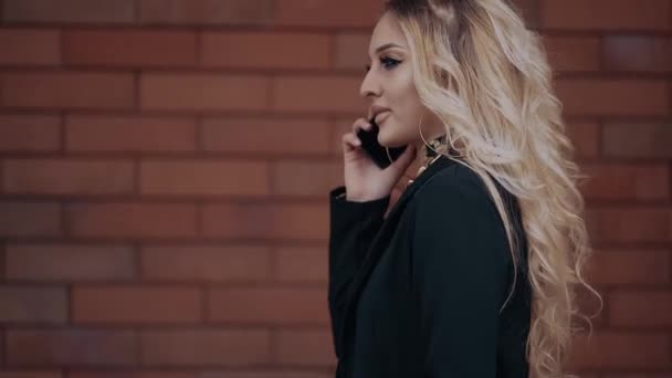 Den unge blondine, karismatisk, filmet i profil, går taler i telefon, samtalen sluttede, og hun lægger på telefonen, går på baggrund af en orange murstensbygning, klædt i en – Stock-video