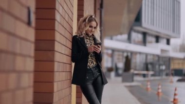 Mutlu genç bayan bir binanın duvarına yaslanıyor, mutlu bir şekilde gülümsüyor ve ileriye bakıyor. Tüm çerçeve, bulanık bir caddenin arka planına karşı. Kadın enerjisi. Güzellik endüstrisi. Sanatsal