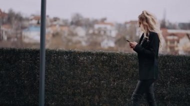 Sarışın, karizmatik kadın, profilde, elinde telefonuyla kaldırımda yürüyor ve gürültülü şehrin arka planına karşı internette geziniyor, ceketli, sert görünümlü...