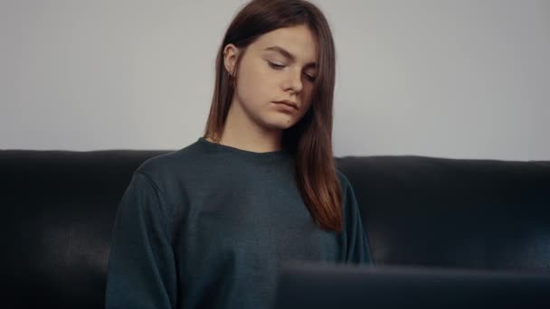 La joven trabajadora, enfocada, escribiendo nuevas ideas para trabajar en su computadora portátil, mira en parte pensativamente, arregla su cabello detrás de su oreja. Vestido con una maleta verde oscuro, sentado en un sofá negro. Concepto 4k — Vídeos de Stock