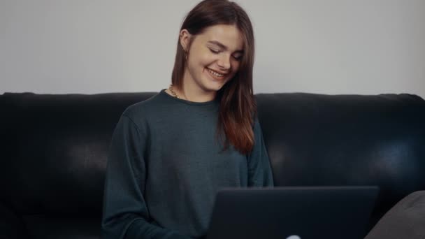 La joven pelirroja pecosa, con una amplia sonrisa, escribió en la pantalla del portátil. Estudiante divertida por su conversación con amigos. Vestido con una maleta verde oscuro, sentado en un sofá negro. Concepto 4k — Vídeos de Stock