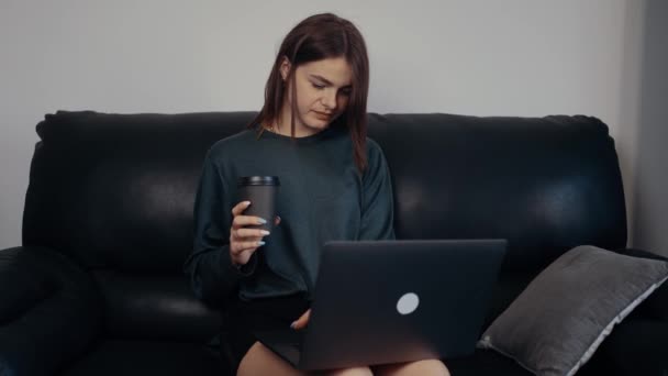 A menina ruiva, empreendedora, de bom humor, trabalha em casa, segura um café na mão, digita um novo projeto em seu laptop. Vestida em uma mala verde escura, sentada em um sofá preto. Conceito 4k — Vídeo de Stock