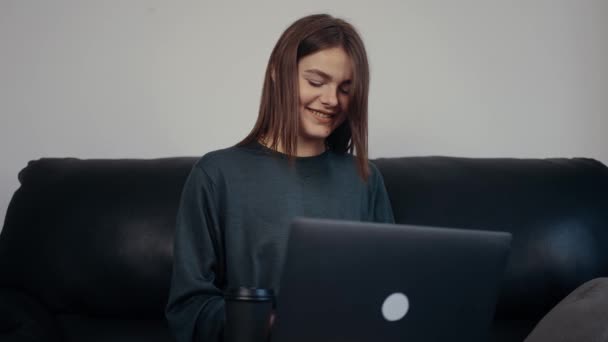 La estudiante alegre con el pelo rojo y la sonrisa ancha escribe el mensaje a sus colegas en su portátil, en las redes sociales. Vestido con una maleta verde oscuro, sentado en un sofá negro. Concepto 4k — Vídeos de Stock