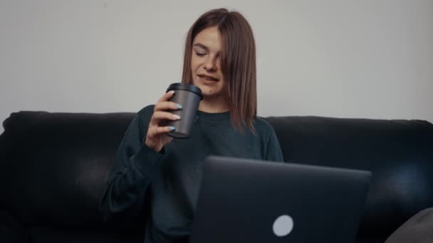 빨간 머리 직장 여성은 집에서 노트북으로 일하며 휴식을 취하고 커피를 마십니다. 짙은 녹색 가방을 걸치고 검은 소파에 앉아 있었다. 4k 개념 — 비디오
