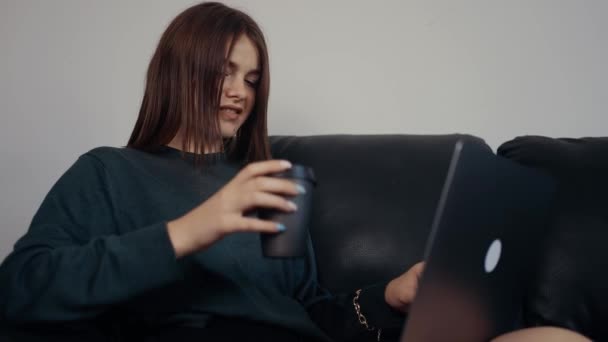 Rudovlasá pihovatá dívka se o víkendu uvolní a povídá si se svými přáteli na notebooku a popíjí tichou kávu. Oblečený do tmavozeleného kufříku, sedící na černé pohovce. Koncept 4k — Stock video