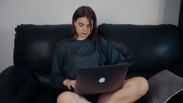 Η νεαρή φοιτήτρια, κοκκινομάλλα με φακίδες, μελετά για τις επόμενες εξετάσεις, κουρασμένη και κάθεται στον καναπέ με το λάπτοπ της στην αγκαλιά της και διαβάζει. Ντυμένη σε μια σκούρα πράσινη βαλίτσα, καθισμένη — Αρχείο Βίντεο