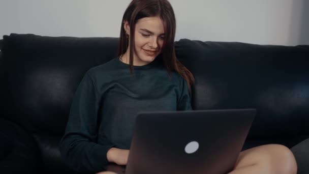 De roodharige met een brede glimlach kijkt naar het laptopscherm en typt iets dat haar amuseert. Gekleed in een donkergroene koffer, zittend op een zwarte bank. Werkconcept. 4k-concept — Stockvideo