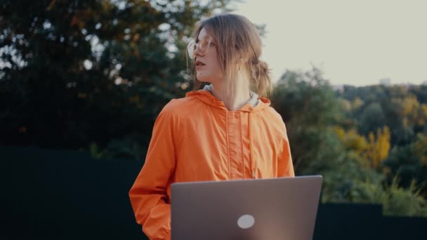 O tânără obosită, cu laptop în brațe, uitându-se în părți și admirând natura din parc. Îmbrăcat într-o bluză portocalie, plasat la intrarea în parc pe fundalul unui apus de soare de basm — Videoclip de stoc