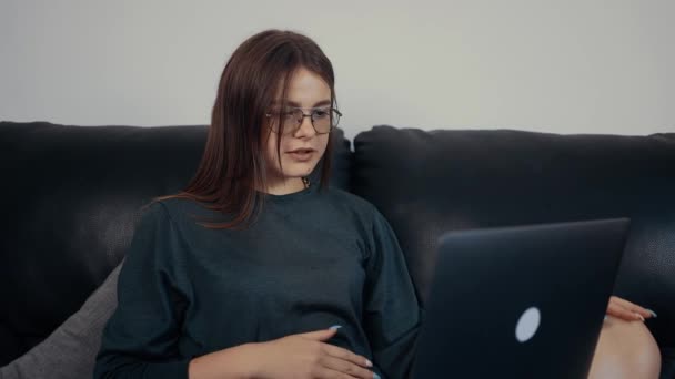 Натхненна молода жінка, червона з веснянками та окулярами, викладає дуже важливий урок онлайн для своїх студентів. Одягнена в темно-зелену блузку, сидить на чорному дивані. Концепція роботи — стокове відео