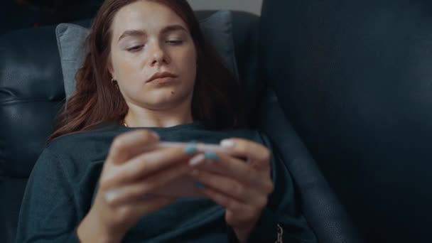 Rödhårig, fräknig, fokuserad på telefonen, liggandes på den svarta soffan hemma. Begreppet samtida kvinna. 4k-konceptet — Stockvideo