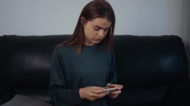 Çilli kızıl saçlı bir kadının portresi telefona odaklanmış, evdeki siyah kanepede oturuyor. Oyuncu konsepti. 4k konsepti