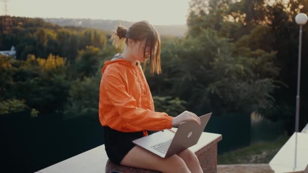 Ambitna młoda kobieta, w okularach, które otwierają laptopa i zaczynają pisać. Ubrany w pomarańczową bluzkę, umieszczony przy wejściu do parku na tle bajkowego zachodu słońca. - Co? — Wideo stockowe