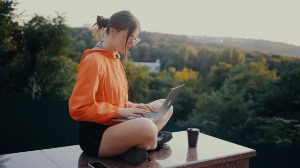这位有灵感、红头发的年轻女子轻拍大学项目，休息一下，渴望身边的咖啡。穿着一件橙色的衬衫，放在公园的入口处，靠着公园。 — 图库视频影像