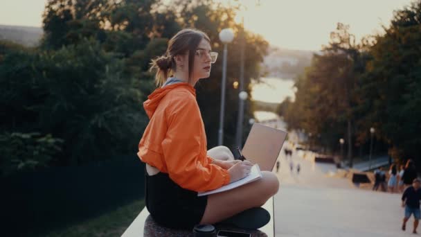 Bedachtzame student die huiswerk maakt in het park, bedachtzaam notities schrijft, met laptop naast haar. Gekleed in een feloranje blouse, haar gevangen in een broodje, gepositioneerd op de parkstrip. 4k-concept — Stockvideo