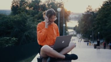 Çalışkan ve iyimser bir kız telefonda konuşur, geniş bir şekilde gülümser, dizüstü bilgisayarın klavyesinde yazarken. Parlak turuncu bir bluz giymişti. Saçları topuz şeklindeydi. Parkta lotus pozisyonundaydı.
