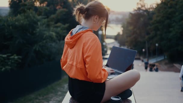 Egy ambiciózus diák a laptopjára írja a főiskolai dolgozatát. Fényes narancssárga blúzba öltözve, zsemlébe akadt hajjal, a park szélén. 4k koncepció — Stock videók