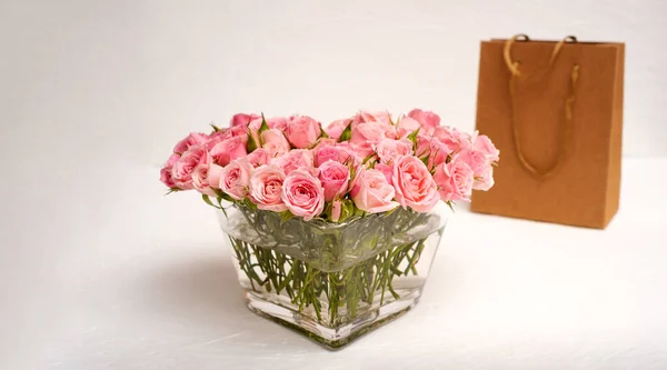 Ramo de rosas rosadas en un jarrón de vidrio con un regalo en una bolsa de artesanía — Foto de Stock