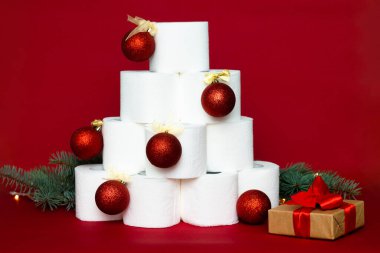 Noel ağacı tuvalet kağıdı rulolarından yapılmıştır, parlak toplarla süslenmiştir, bir hediyenin yanında ve kırmızı arka planda köknar dalları vardır.