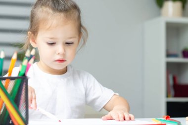 Beyaz tişörtlü şirin küçük kız 2-4 renkli keçeli kalemler ve kalemlerle evde çizim yapıyor.