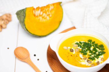 Kremalı sarı balkabağı kremalı çorba, zencefil, çiğ balkabağı ve ahşap kaşık yakınındaki beyaz bir masada otlar ve tohumlar..