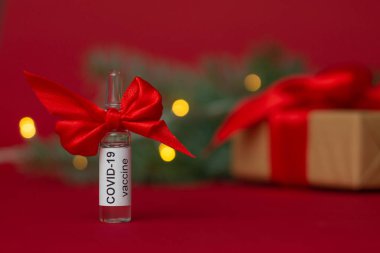 Bir ampul COVID-19 Coronavirus aşısı kırmızı arka planda ışıkları olan bir Noel ağacı dalının yanında hediye olarak duruyor.