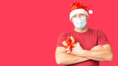Noel Baba şapkalı ve tıbbi maskeli bir adam kırmızı arka planda bir Noel hediyesi tutuyor..
