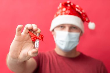Noel Baba şapkası ve tıbbi maske takan bir adam kırmızı arka planda COVID-19 aşısını tutuyor..