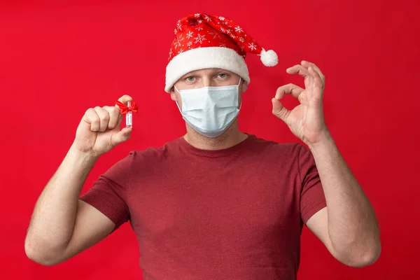 Ένας άντρας με καπέλο Αϊ Βασίλη και ιατρική μάσκα κρατάει το εμβόλιο COVID-19 και δείχνει την πινακίδα ΟΚ σε κόκκινο φόντο.. — Φωτογραφία Αρχείου