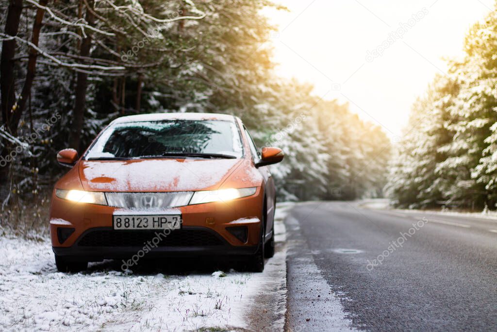 03.12.2020 Minsk, Belarus: orange Honda Civic 2008 with headlights on on asphalt road in a snowy pine forest with sun rays