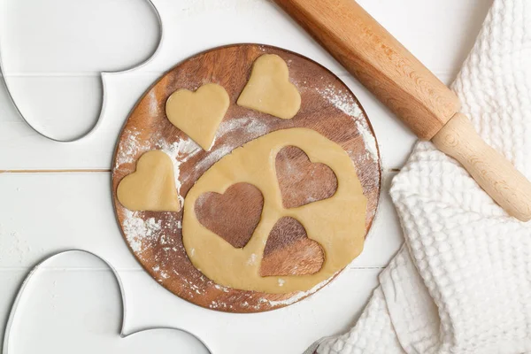 A heart shaped cutout mold and dough cut cookies next to a towel and wooden spoons and rolling pin. Flat lay.