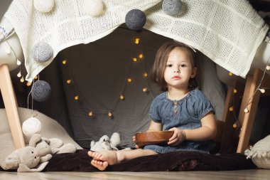 2-4 numaralı kız sandalye ve battaniye kulübesinde oturuyor. Çocuk evde oyuncaklarla oynuyor