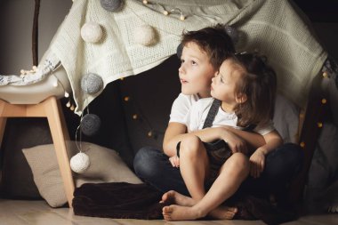 Kardeşler sandalye ve battaniye kulübesinde sarılırlar. Erkek ve kız kardeş evde oynuyor.