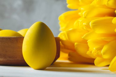 Parlak sarı lale buketi ve Paskalya yumurtası, gri arka planda beyaz ahşap yüzeyde duruyor.