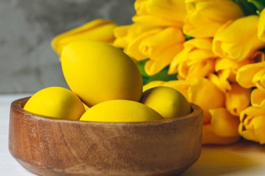 Paskalya yumurtaları ahşap kasede ve parlak sarı lalelerden oluşan buket beyaz ahşap zemin üzerinde gri arka planda.