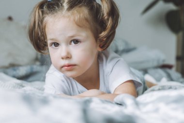 Koyu renk saçlı küçük bir kız. 2-4, iki at kuyruklu. Yatağında uzanmış, çerçeveye bakıyor.