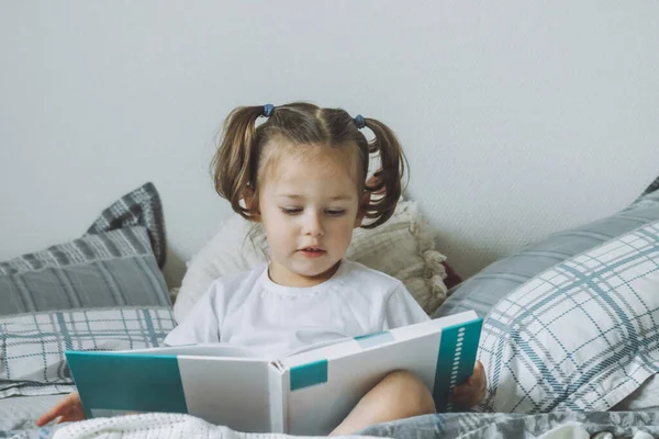 Klein donkerharig meisje 2-4 met twee paardenstaarten zit op bed met kussens en leest boek — Stockfoto