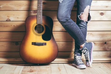 Altı telli akustik sarı-kahverengi gitar ve yırtık kot pantolonlu kadın bacakları, ahşap duvarın yanında.