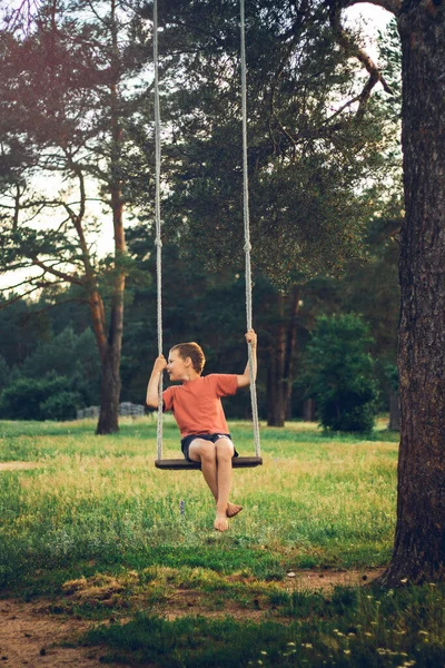 Barefoot jongen 7-10 zittend op touw schommel op het platteland tussen pijnbomen in stralen van ondergaande zon. Verticaal — Stockfoto