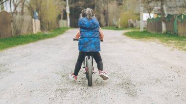 Mavi yelekli ve siyah taytlı çocuk toprak yolda dengeli bisiklete biniyor.