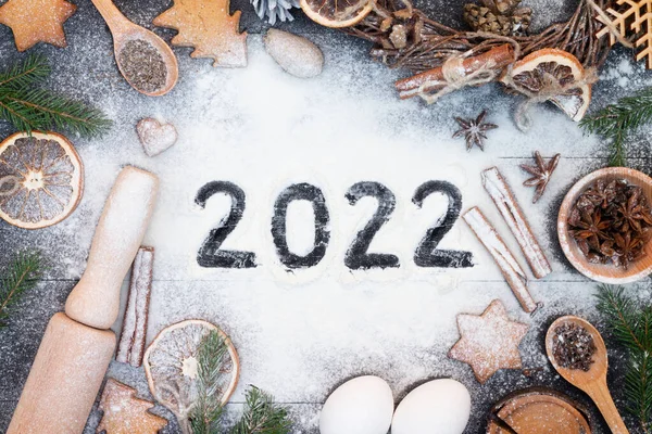 Happy New Year 2022 written on flour. Christmas tree branches, gingerbread cookies, spices and baking supplies on black wood background. Christmas, New Year greeting card