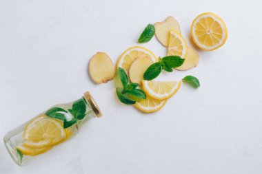 İçinde limonata ve yemek pişirmek için malzemeler olan cam şişe. Zencefil, limon, beyaz yüzeyde nane. Düz yatış, üst görünüm