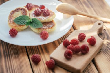 Lezzetli peynirli kekler ya da pudra şekerli krepler ahşap masaya ahududu ve nane yaprağı serpiştirilmiş.