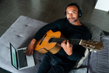 Evde gitar çalıp dizüstü bilgisayar kullanan rahat bir adam.