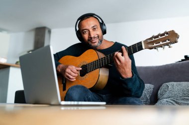 40 yaşında bir adam evde gitar çalıyor ve dizüstü bilgisayar kullanıyor.