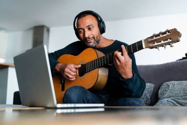 Evde gitar çalıp dizüstü bilgisayar kullanan rahat bir adam.