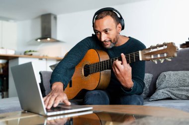 Evde gitar çalıp dizüstü bilgisayar kullanan rahat bir adam.