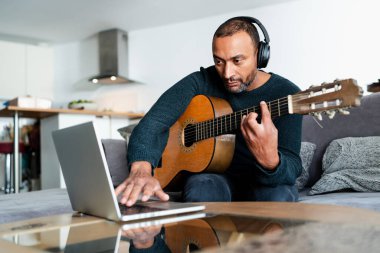 Siyah adam evde gitar çalıyor ve dizüstü bilgisayar kullanıyor.