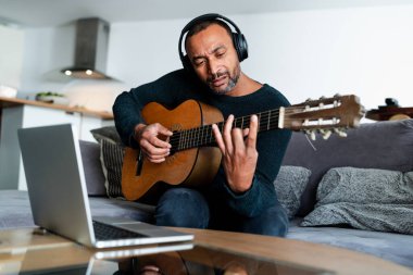 Evde gitar çalıp dizüstü bilgisayar kullanan rahat bir adam.